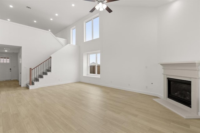 unfurnished living room with ceiling fan, a glass covered fireplace, a high ceiling, stairs, and light wood-style flooring