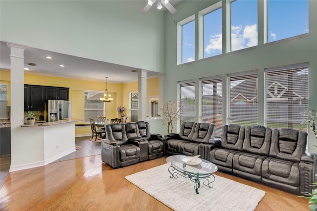 living room with ornate columns, a high ceiling, ceiling fan with notable chandelier, and light wood-type flooring