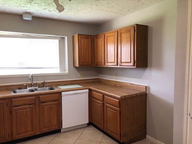 kitchen with a textured ceiling, white dishwasher, brown cabinets, a sink, and light tile patterned flooring