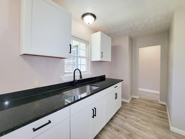 kitchen with a sink, a textured ceiling, dark stone counters, white cabinetry, and light wood-type flooring