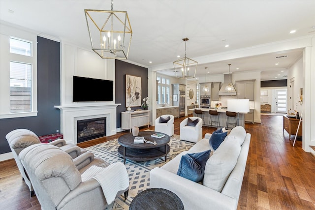 living area with crown molding, a glass covered fireplace, dark wood-style floors, and an inviting chandelier