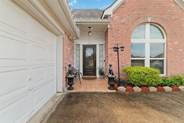 view of exterior entry with brick siding, a garage, roof with shingles, and driveway