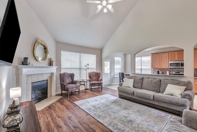 living room featuring a towering ceiling, light wood-type flooring, arched walkways, a tiled fireplace, and a ceiling fan