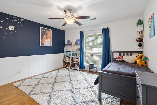 bedroom featuring a ceiling fan, visible vents, wood finished floors, and a wainscoted wall