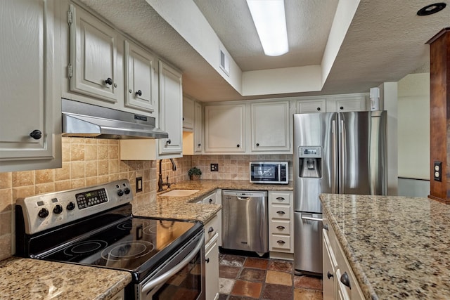 kitchen featuring appliances with stainless steel finishes, a sink, stone finish floor, backsplash, and under cabinet range hood