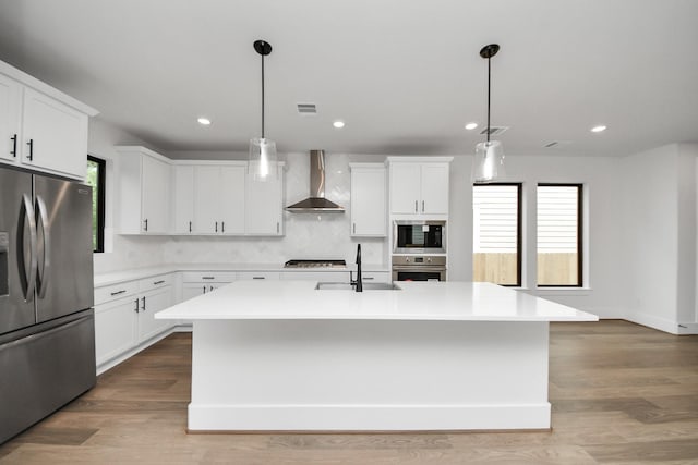 kitchen featuring a kitchen island with sink, wall chimney range hood, light countertops, a sink, and stainless steel appliances