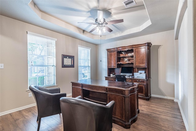 office space with visible vents, wood finished floors, a healthy amount of sunlight, a tray ceiling, and a ceiling fan