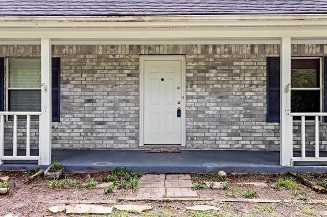 doorway to property with covered porch, a shingled roof, and brick siding