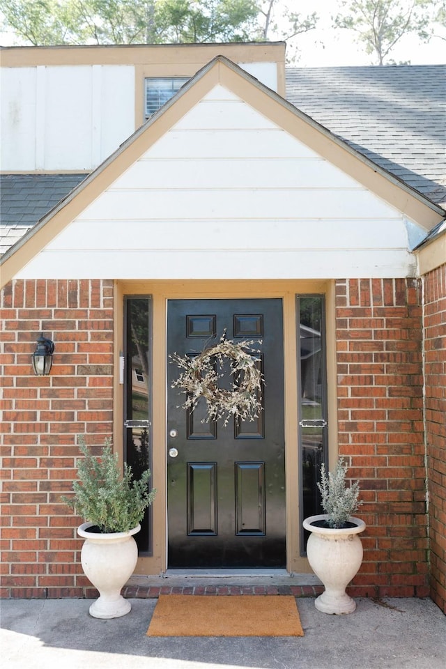 view of exterior entry with a shingled roof and brick siding