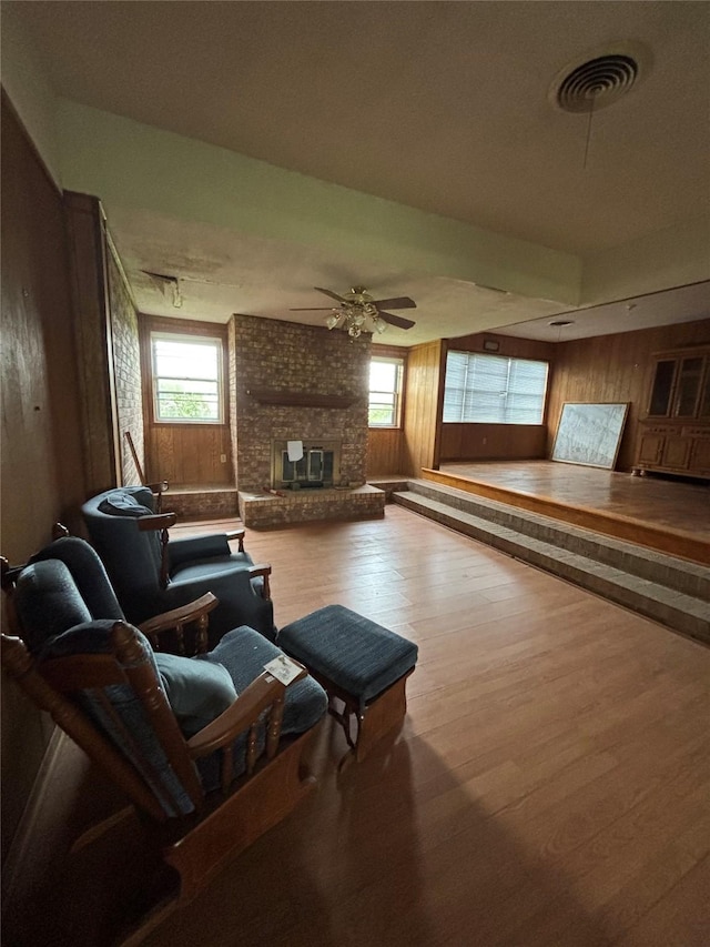 living area featuring a brick fireplace, wood walls, a ceiling fan, wood finished floors, and visible vents