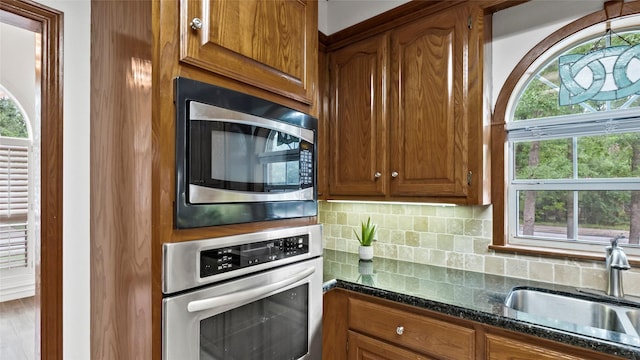 kitchen featuring brown cabinets, appliances with stainless steel finishes, a sink, dark stone counters, and tasteful backsplash