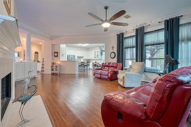 living area featuring ceiling fan, visible vents, crown molding, wood finished floors, and ornate columns