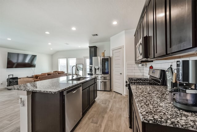 kitchen with appliances with stainless steel finishes, a sink, light wood-style flooring, dark stone countertops, and tasteful backsplash