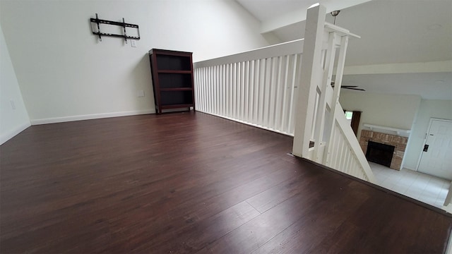 unfurnished room featuring a brick fireplace, wood finished floors, baseboards, a ceiling fan, and vaulted ceiling