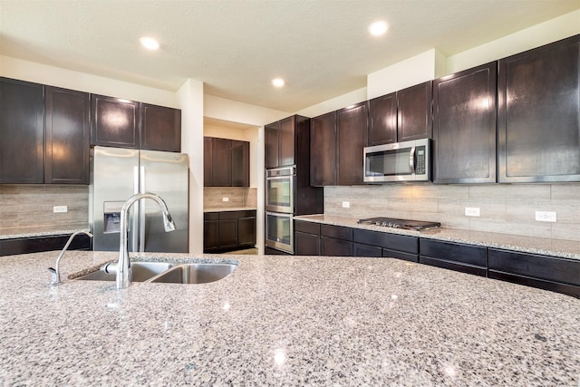 kitchen featuring stainless steel appliances, a sink, light stone countertops, dark brown cabinetry, and recessed lighting