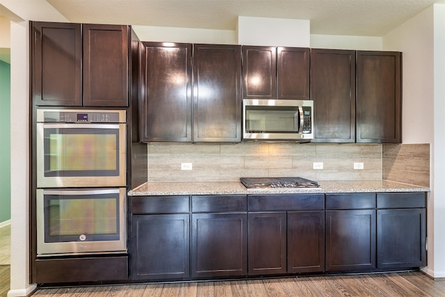 kitchen with appliances with stainless steel finishes, wood finished floors, decorative backsplash, dark brown cabinets, and light stone countertops