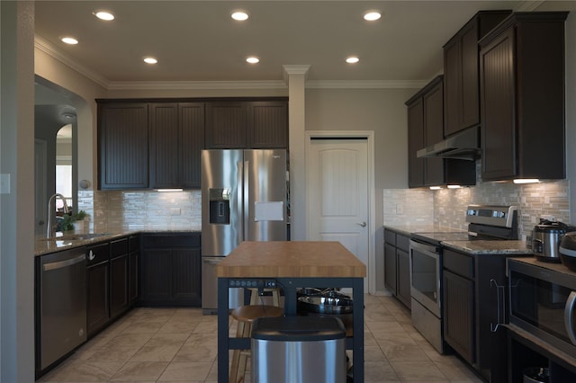 kitchen featuring crown molding, a sink, under cabinet range hood, dark stone counters, and stainless steel appliances