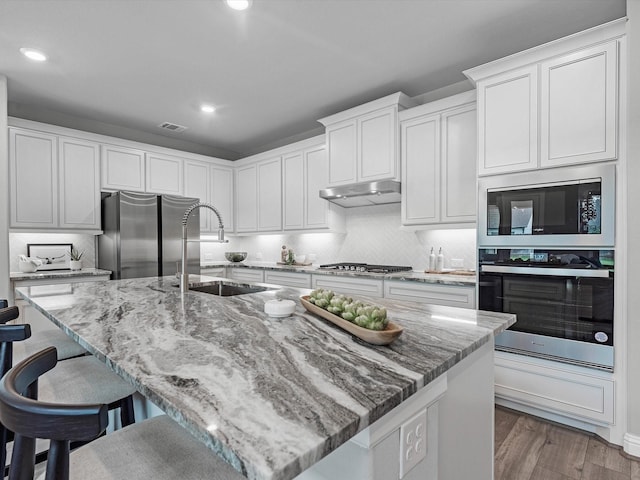 16926 Grayson Woods Ln, Humble, TX 77346 - kitchen with a kitchen island with sink, under cabinet range hood, light stone counters, a sink, and appliances with stainless steel finishes
