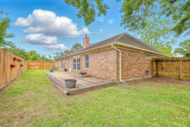 rear view of property featuring a wooden deck, a fenced backyard, a chimney, brick siding, and a lawn