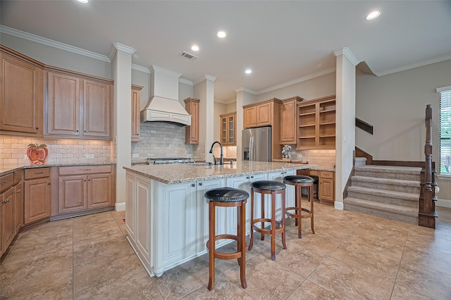 kitchen with open shelves, custom exhaust hood, light stone countertops, a center island with sink, and stainless steel fridge