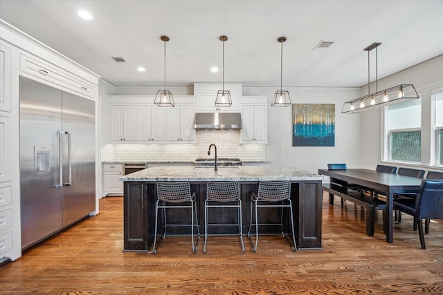 kitchen featuring visible vents, wood finished floors, built in refrigerator, under cabinet range hood, and white cabinetry