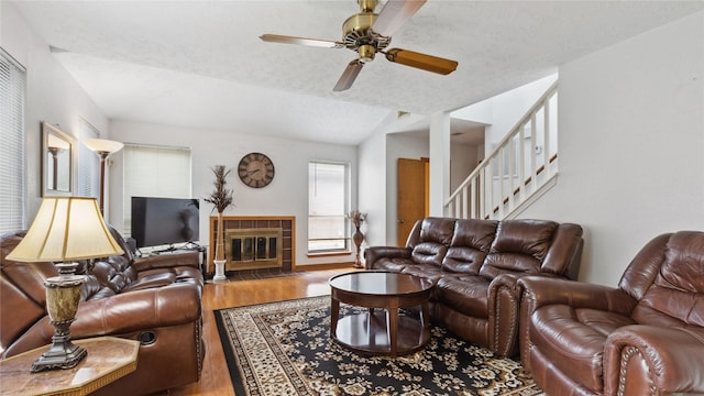 living area with wood finished floors, stairway, a tiled fireplace, a ceiling fan, and vaulted ceiling
