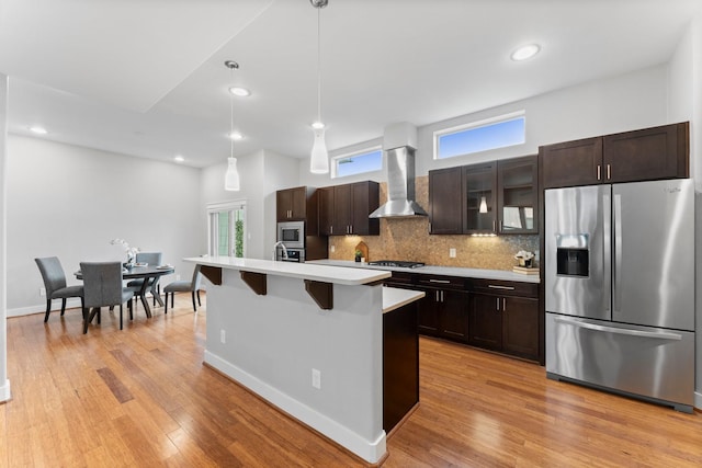 kitchen with wall chimney range hood, dark brown cabinetry, light countertops, stainless steel appliances, and backsplash