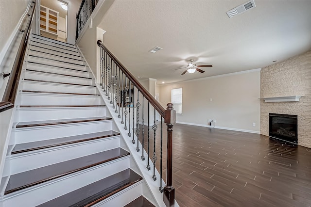 staircase featuring a stone fireplace, a ceiling fan, baseboards, wood finished floors, and visible vents