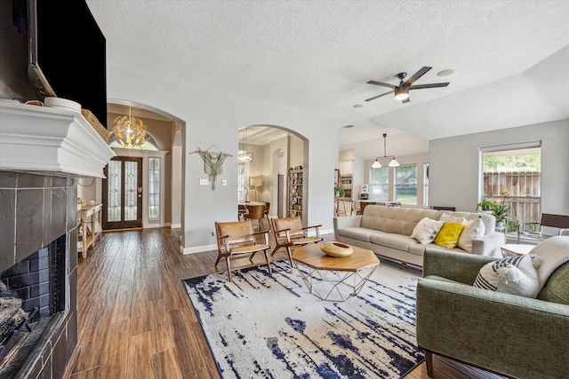 living area with ceiling fan with notable chandelier, a fireplace, a textured ceiling, arched walkways, and dark wood-style floors