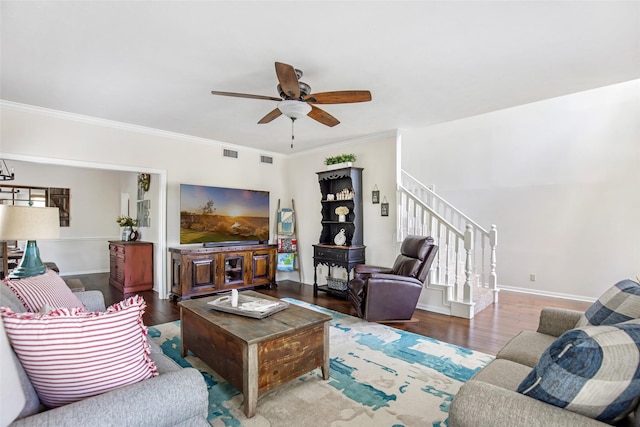 living room featuring a ceiling fan, visible vents, crown molding, wood finished floors, and stairs