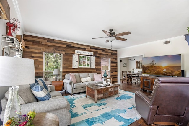 living room with wooden walls, ceiling fan, wood finished floors, visible vents, and ornamental molding