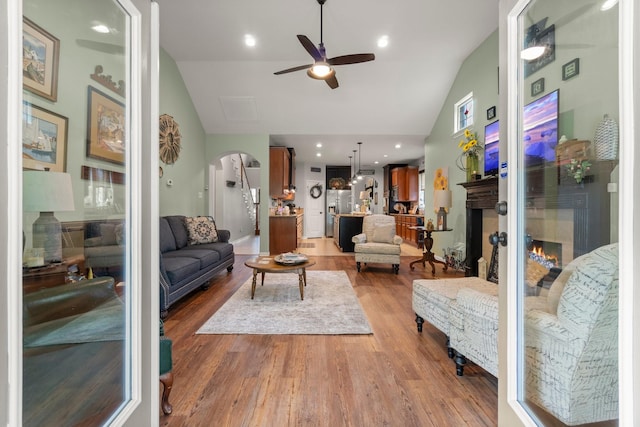 living room featuring a ceiling fan, arched walkways, wood finished floors, and a warm lit fireplace