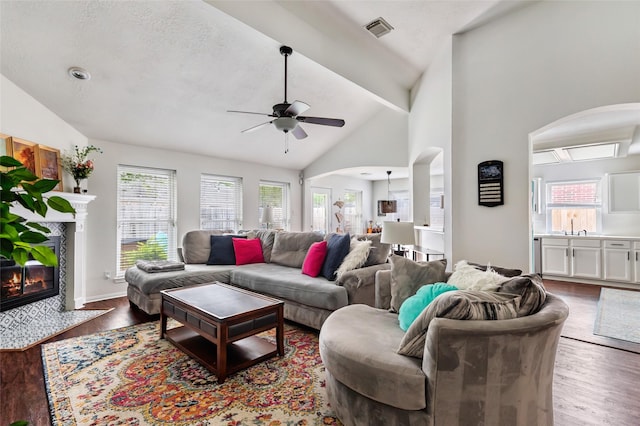 living room featuring arched walkways, a ceiling fan, wood finished floors, a tiled fireplace, and high vaulted ceiling