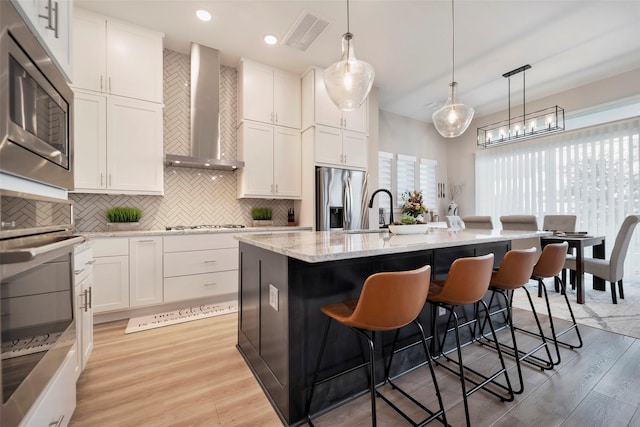 kitchen featuring wall chimney range hood, light wood-style floors, a breakfast bar area, stainless steel appliances, and visible vents