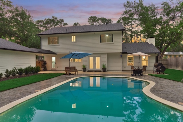 back of house at dusk with french doors, a patio, fence, a chimney, and roof with shingles