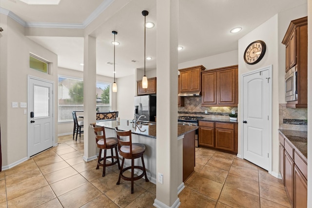 kitchen featuring brown cabinets, appliances with stainless steel finishes, a breakfast bar area, backsplash, and a center island with sink