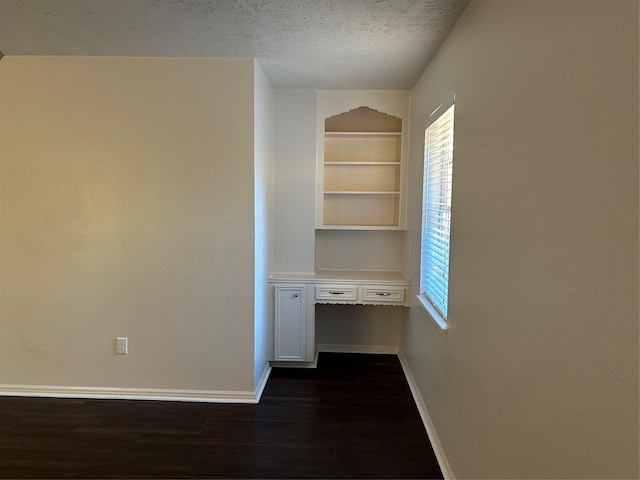 spare room with baseboards, built in study area, dark wood-type flooring, a textured ceiling, and built in shelves