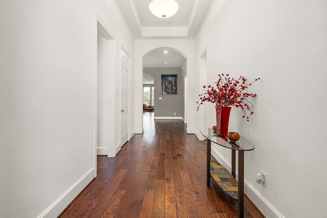 hallway featuring arched walkways, dark wood-style flooring, baseboards, and a raised ceiling