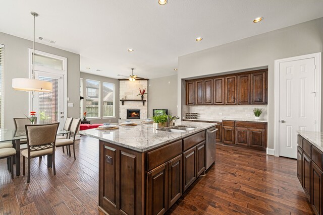 kitchen featuring a sink, a stone fireplace, stainless steel dishwasher, dark wood-style flooring, and ceiling fan