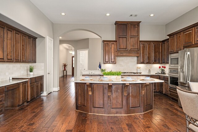 kitchen with stainless steel appliances, arched walkways, backsplash, a kitchen island, and dark brown cabinets