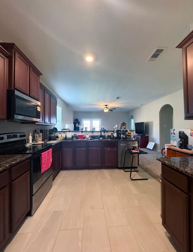 kitchen with appliances with stainless steel finishes, open floor plan, a ceiling fan, arched walkways, and dark stone countertops