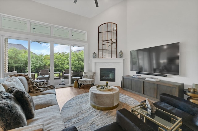 living room featuring wood finished floors, a towering ceiling, a healthy amount of sunlight, a ceiling fan, and a glass covered fireplace