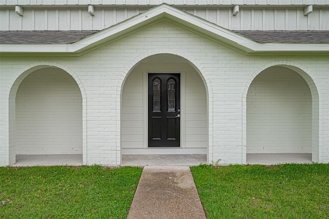 doorway to property featuring a yard, roof with shingles, brick siding, and board and batten siding