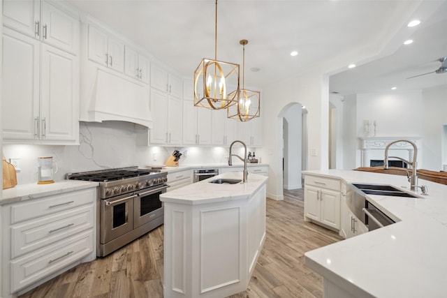 kitchen with a sink, a center island with sink, arched walkways, appliances with stainless steel finishes, and custom range hood