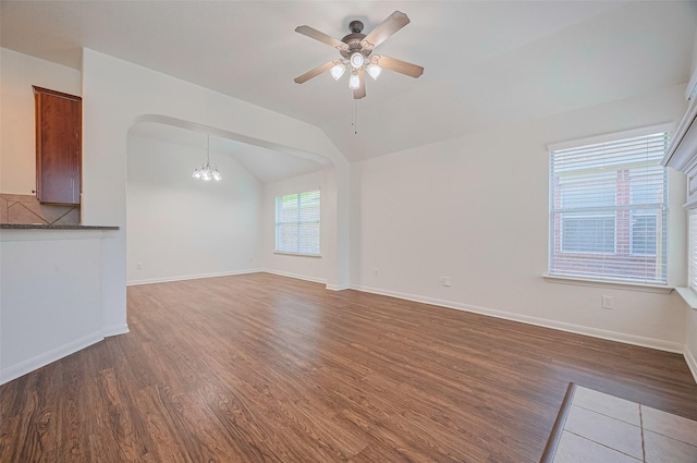 unfurnished living room with lofted ceiling, ceiling fan with notable chandelier, wood finished floors, arched walkways, and baseboards