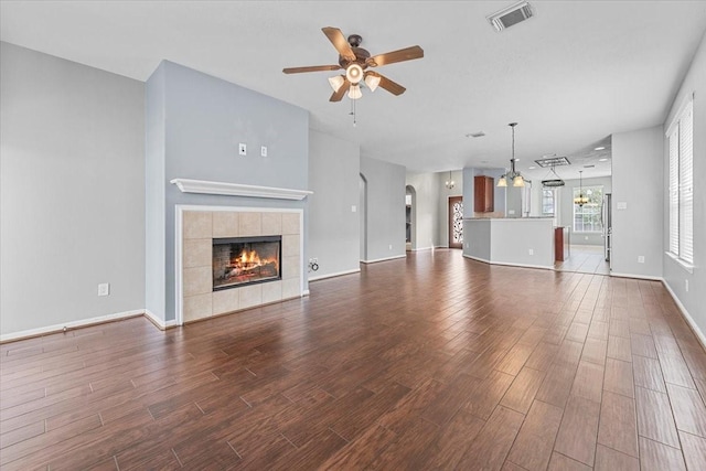 unfurnished living room with baseboards, dark wood-style flooring, a tiled fireplace, visible vents, and ceiling fan with notable chandelier
