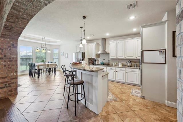 kitchen with wall chimney range hood, decorative light fixtures, visible vents, a kitchen bar, and white cabinets