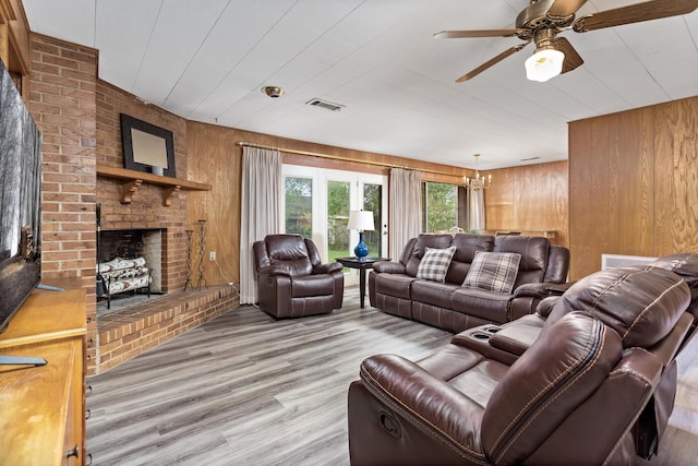 living room featuring light wood finished floors, ceiling fan with notable chandelier, a fireplace, visible vents, and wooden walls