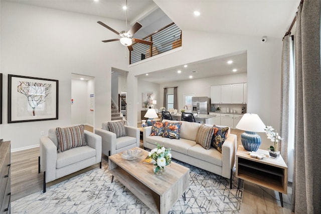 living area with high vaulted ceiling, stairs, light wood-type flooring, and a ceiling fan