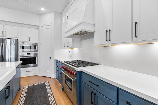 203 S Empress Green Pl, Montgomery, TX 77316 - kitchen featuring appliances with stainless steel finishes, white cabinetry, blue cabinetry, custom range hood, and light wood-style floors